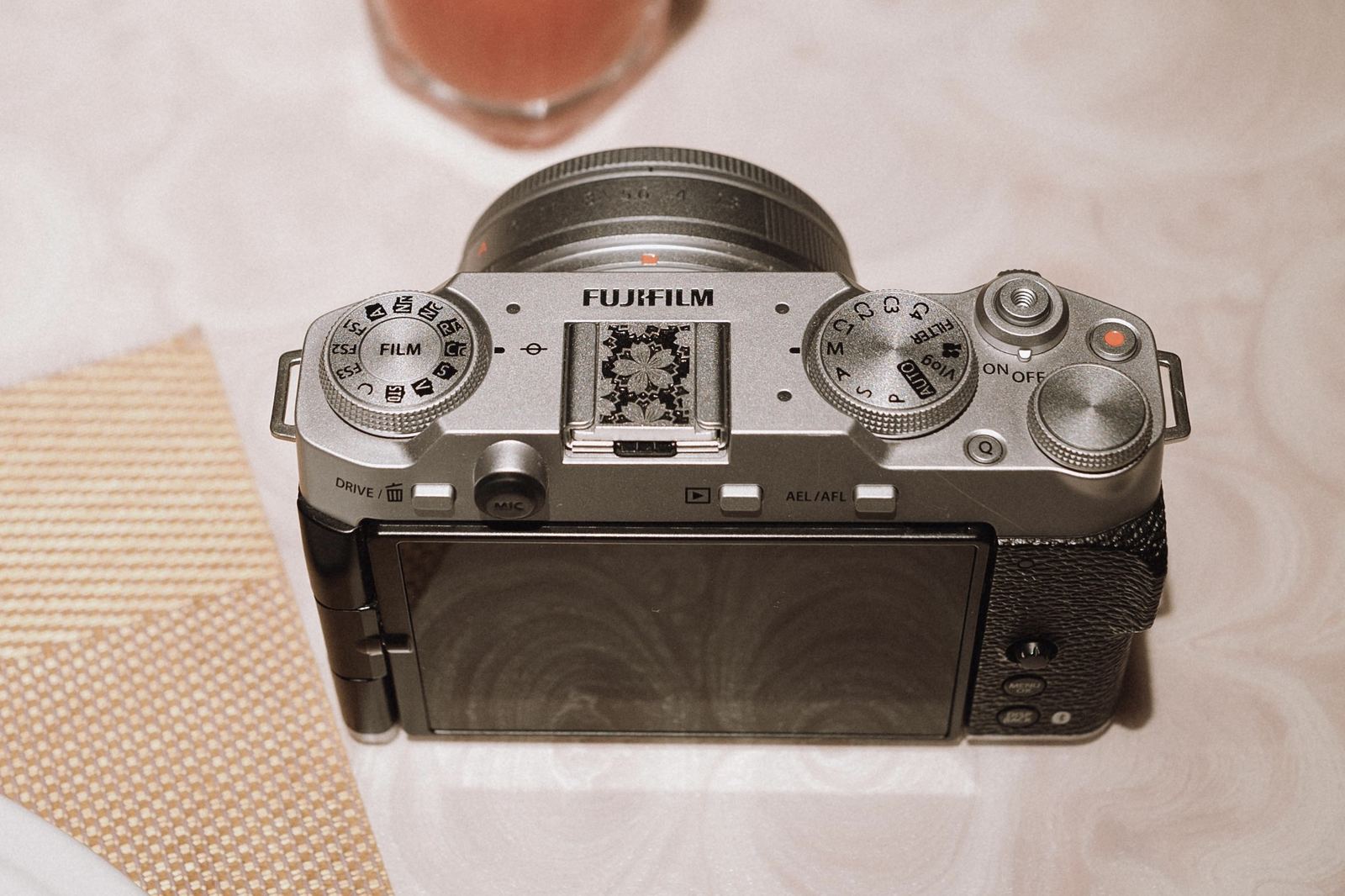 A silver Fujifilm X-M5 on a restaurant table, seen from above, showing the film simulation dial, the mode dial with its Vlog position, the MIC button and the vari-angle screen folded out.