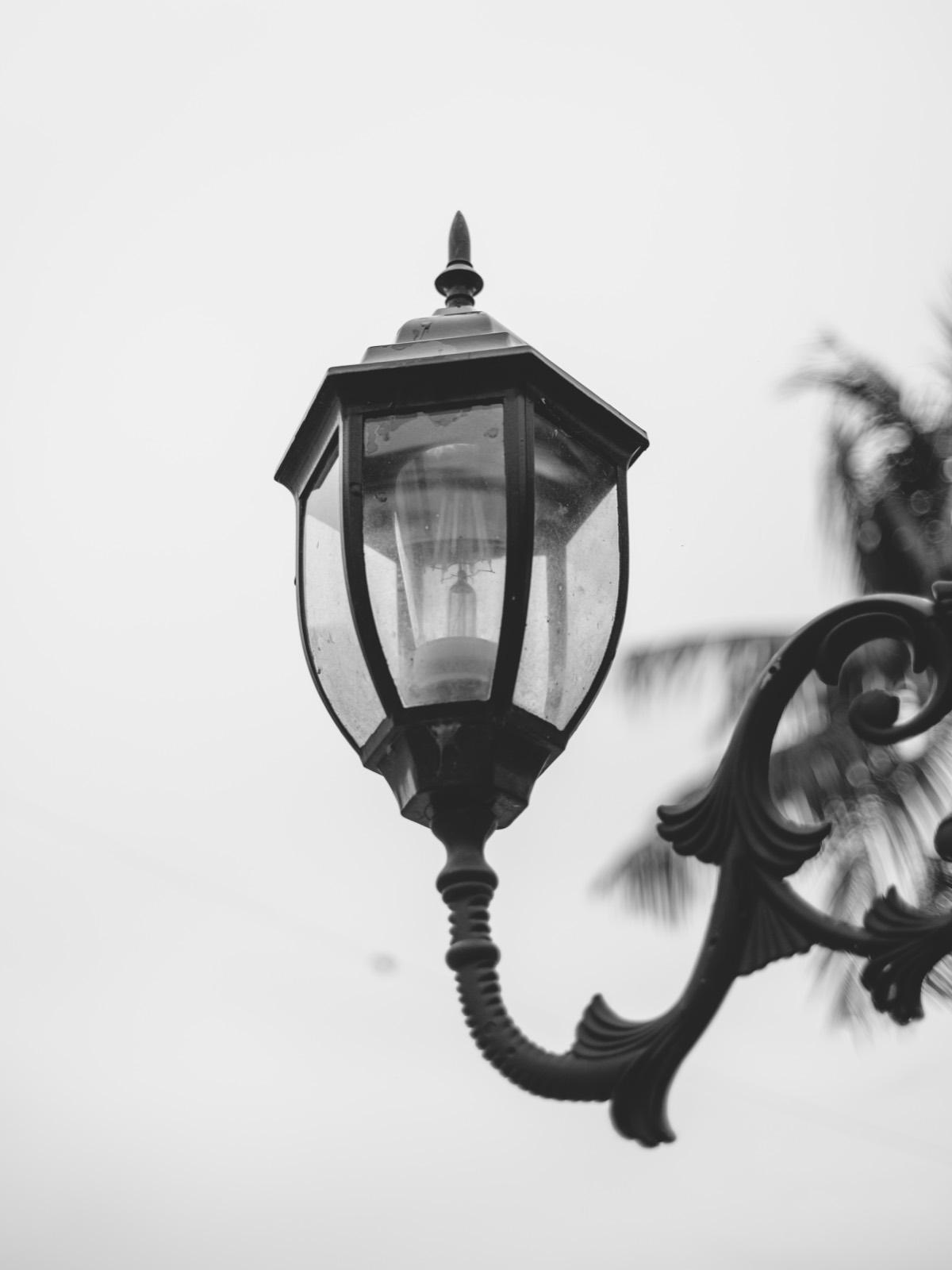 Black-and-white photo of an ornate cast-iron street lamp with a filament bulb against a plain bright sky, shot wide open on the Voigtlander 27mm f/2 on a Fujifilm X-E5.