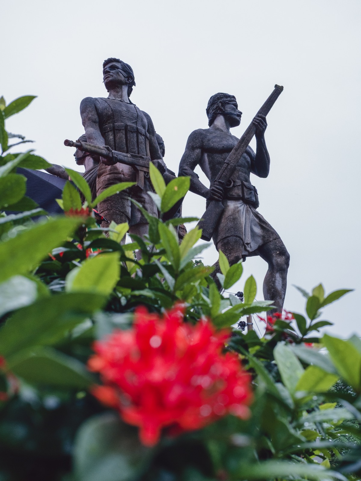 A bronze monument of freedom fighters holding rifles seen from below through blurred red ixora flowers and green leaves, shot on a Fujifilm X-E5 with the Voigtlander 27mm at f/8.
