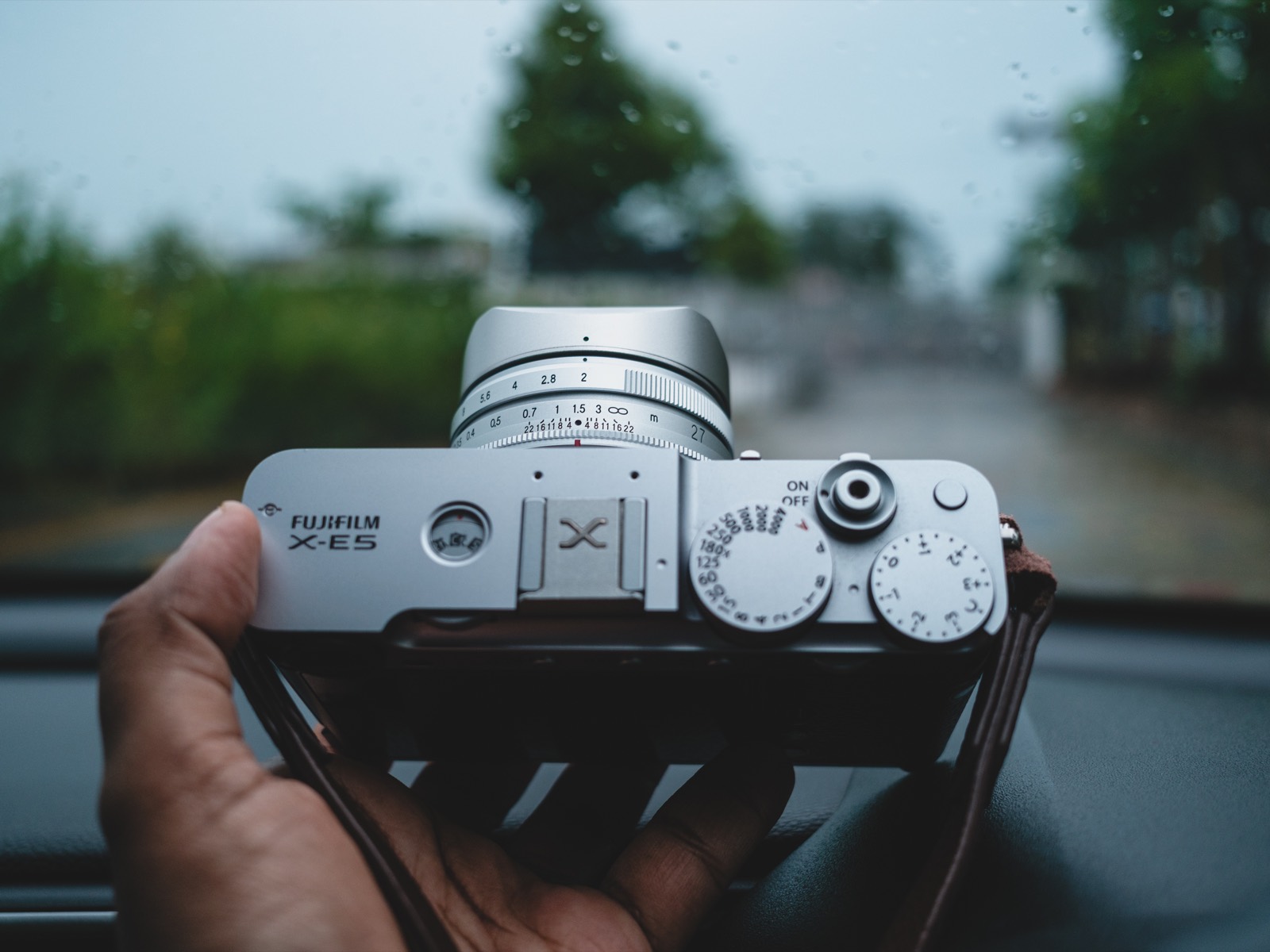 A silver Fujifilm X-E5 held in one hand inside a car, fitted with a silver Voigtlander Ultron 27mm f/2 lens and a square metal lens hood, rain-streaked windscreen and green trees behind.